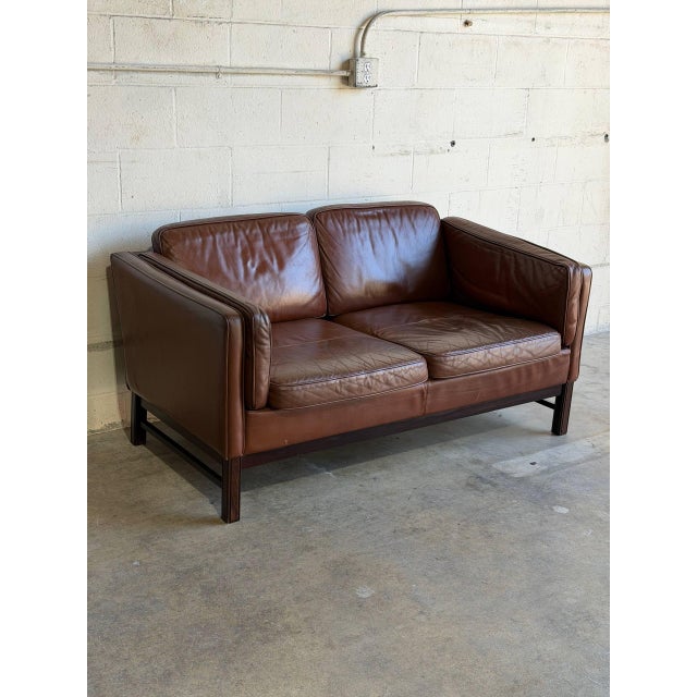 This two-seater sofa features plush brown leather cushions set within a structured rectilinear frame. Its low profile...