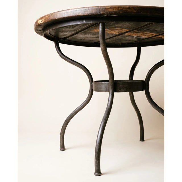 Round dining table featuring a substantial wood plank top paired with a curved wrought iron base. The tabletop has a warm,...