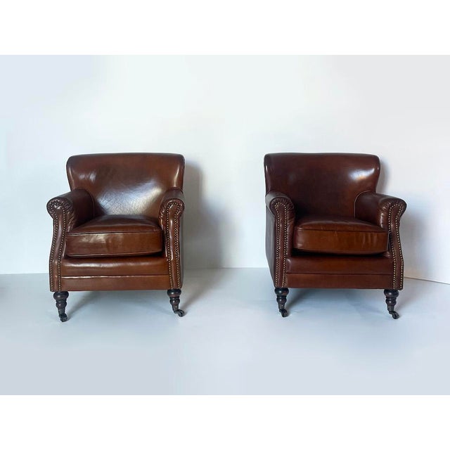 Elegant club-style armchairs upholstered in brown leather with a semi-gloss finish. They feature rounded armrests with...