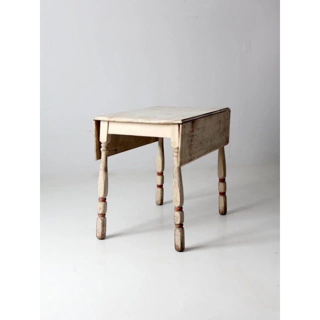 This is an antique drop-leaf kitchen table. Cream paint colors the wooden table with red accents on the turned legs. The...