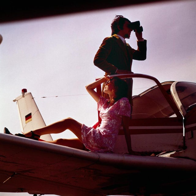 This photograph from 1972 captures a stylish, vibrant scene featuring two people on a small airplane. A young woman,...