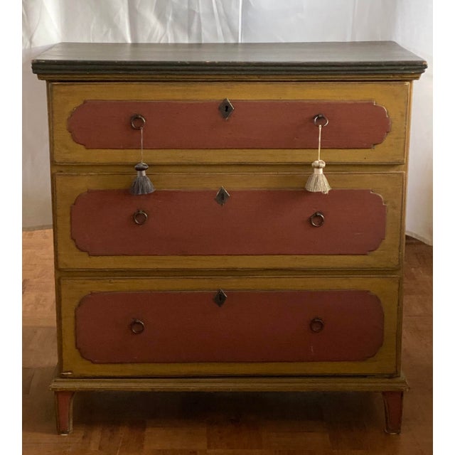 A chest of drawers in cream and old pink with a dark top. There are three drawers, each with two metal rings for opening...
