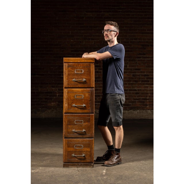 Tall oak filing cabinet featuring four drawers with inset panel faces, iron pulls, and label slots. Each drawer is built...