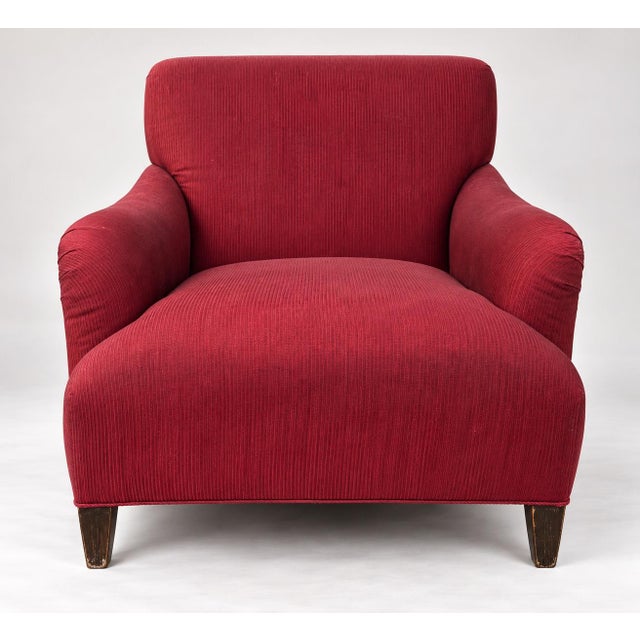 An upholstered armchair featuring low flared arms, a deep cushioned seat, and a tight backrest covered in a deep red...