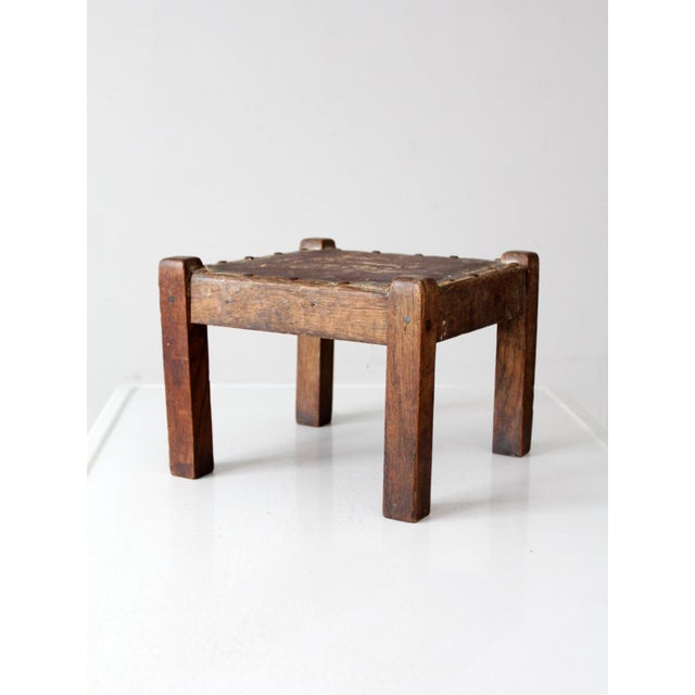 Turn of the century wooden footstool with a compact square form and a time-softened upholstered top secured with hand-set...