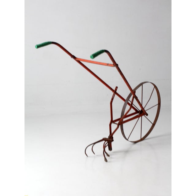 Antique single-wheel garden cultivator with a large spoked iron wheel, curved cultivating tines, and long metal handles....