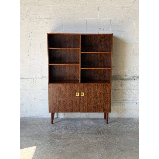 This architectural shelving unit features upper open shelves and lower cabinet doors. Elevated on round tapered legs, it...