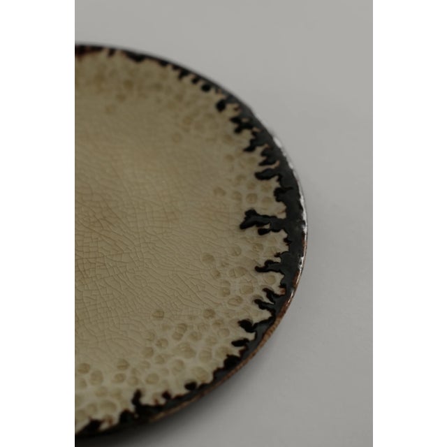 Wheel-thrown plate with a gently curved form, finished in a crackled cream glaze with a dark, irregular edge. Subtle...