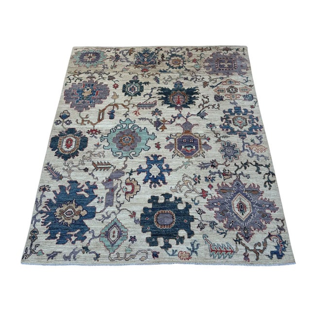Oversized flowers in denim blue, deep teal, lavender, and pale aqua spread across the ivory field of this handmade Oushak...