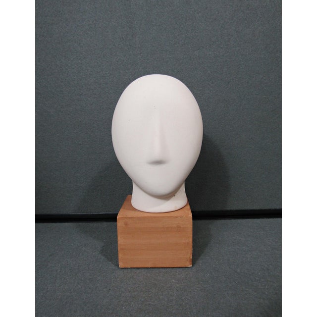 A refined Cycladic-style head sculpture rendered in matte bisque ceramic and presented on a simple wooden plinth. The...