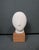 A refined Cycladic-style head sculpture rendered in matte bisque ceramic and presented on a simple wooden plinth. The...