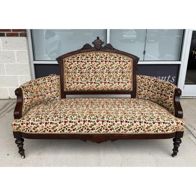 This antique Eastlake settee features a carved walnut frame with an arched upholstered back, shaped arms, turned front...