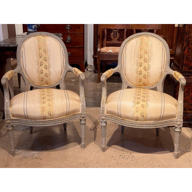 Pair of Louis XVI painted fauteuils with carved oval backs, padded arms on leaf-carved supports and guilloche seat rails,...
