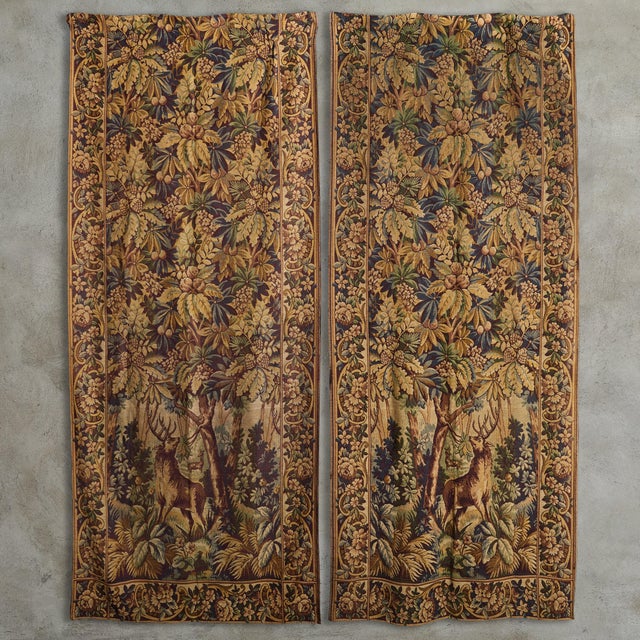 A tall 1920s French tapestry featuring a lush woodland scene with foliate and deer motifs in brown and taupe hues. Time-...