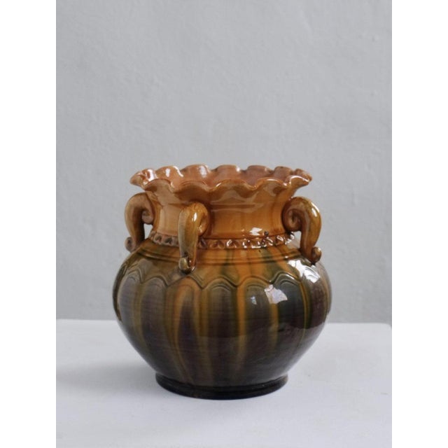 Ornate ceramic vase from Latvia, featuring curled handles and a scalloped rim. The glossy glaze transitions from warm...