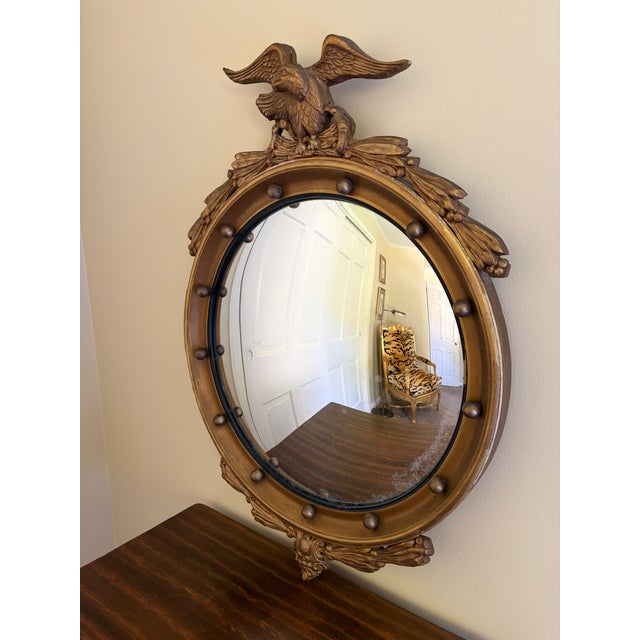 A Federal-style giltwood convex mirror crowned with a carved eagle, its wings outstretched above a laurel motif—an...