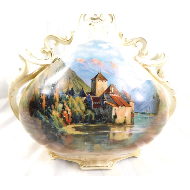 This is an off white Royal Bayreuth vase or jug with a scene of a building by a lake on one side and a sad little green...