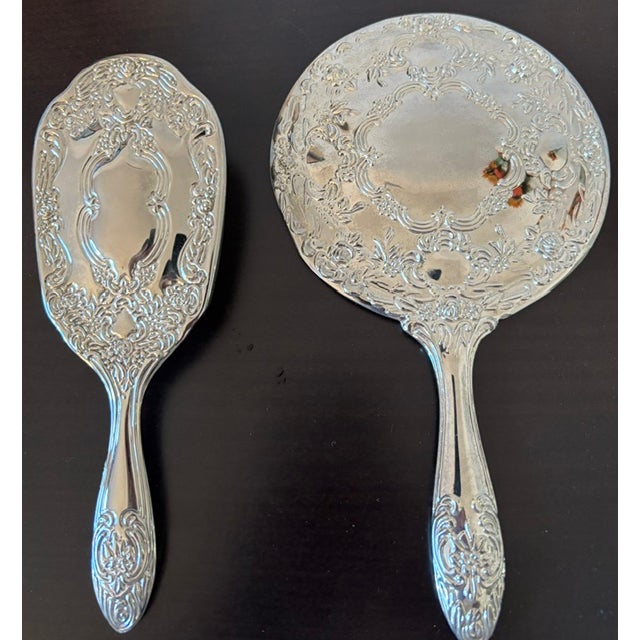 A decorative vintage vanity set featuring a large circular hand mirror and coordinating hairbrush, both embellished with...