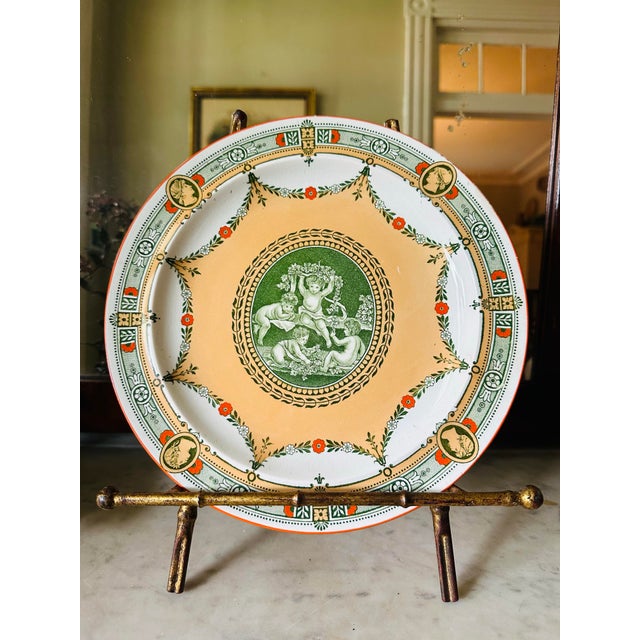A beautiful antique transferware plate featuring a classical cherub scene at the center, surrounded by an elegant...
