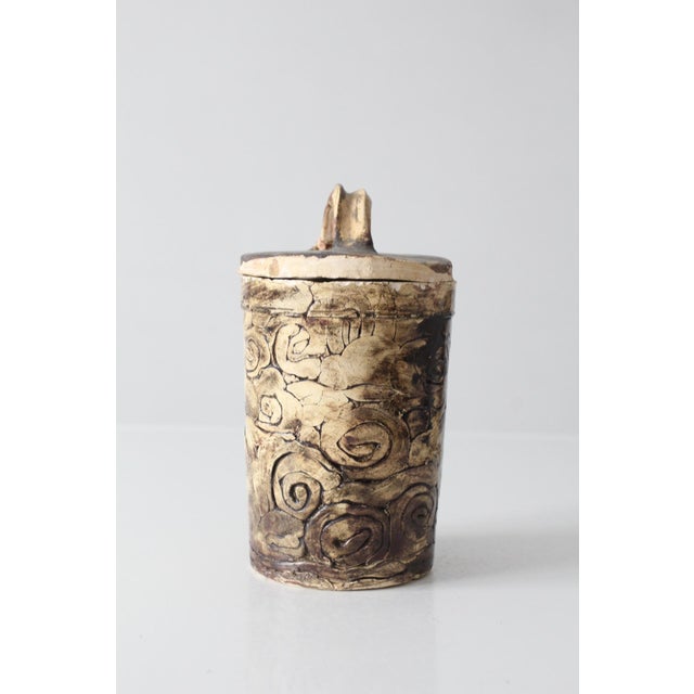 A striking vintage studio pottery jar featuring deeply carved decoration, an earthy palette, and an arched handle rising...