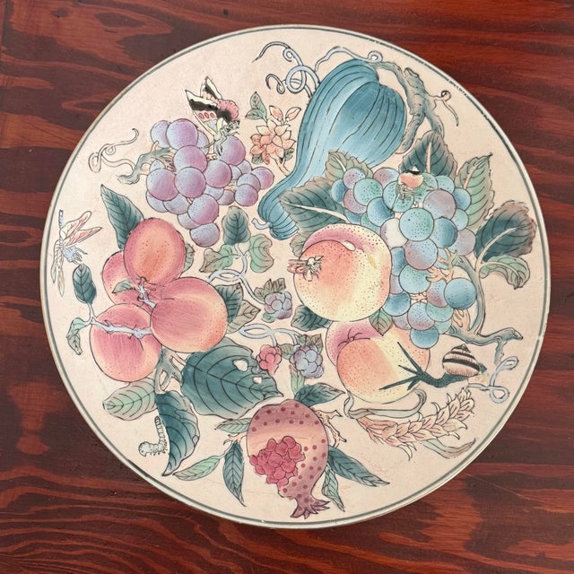 A hand-painted porcelain plate with peaches, grapes, pomegranates, and a small bird among leaves. The colors are soft...