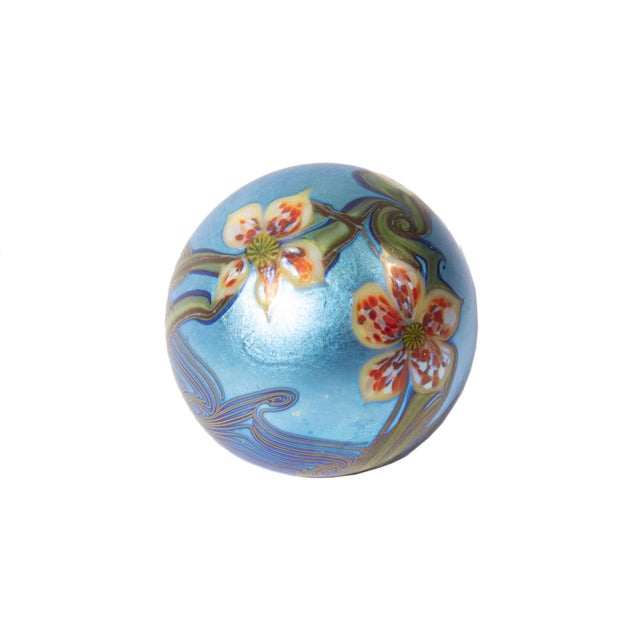 A 1977 iridescent art glass paperweight by Orient and Flume. This spherical paperweight features orange flowers against a...