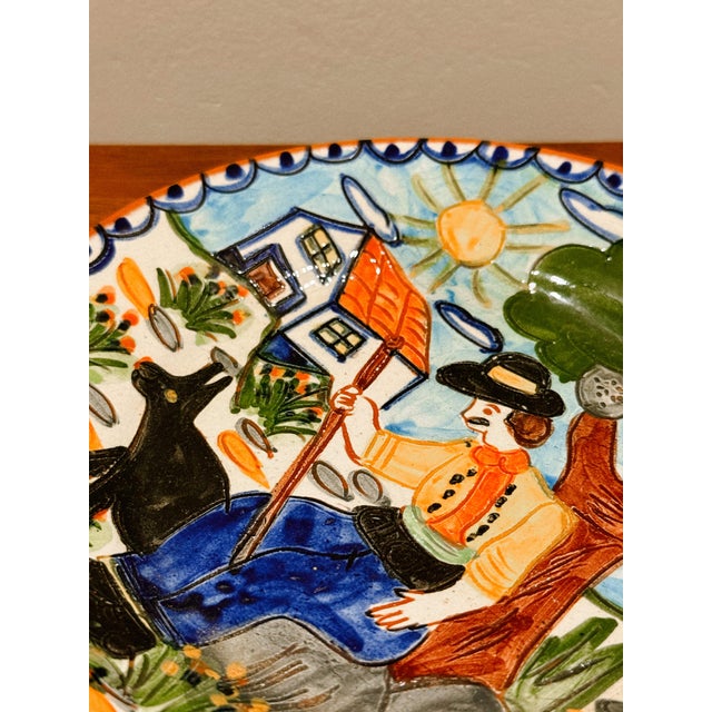 Decorative glazed ceramic plate, hand-painted with a rural figurative scene, depicting a seated figure under a tree in a...