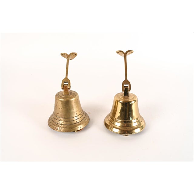 A pair of brass wall-mounted bells with a flared, stepped bell body and a long hinged bracket ending in a forked, two-hole...