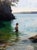 "Between Sea and Sky" is an oil on panel painting by Jon Doran, completed in 2026. In a sheltered cove, a woman in a dark...