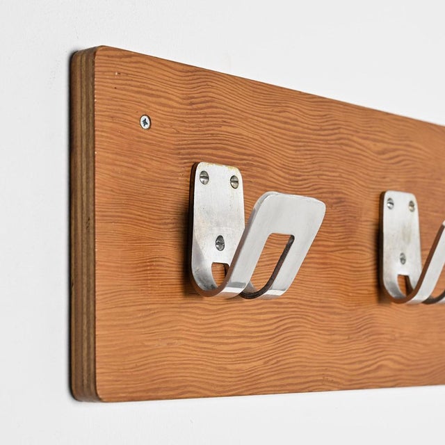 Wall-mounted coat rack composed of a plywood panel veneered in light mahogany, to which four aluminum hooks are attached....