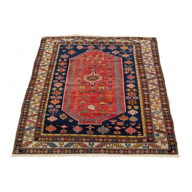This wool rug features a tribal pattern with yellow, beige, and blue accents on a red background, inspired by Persian...