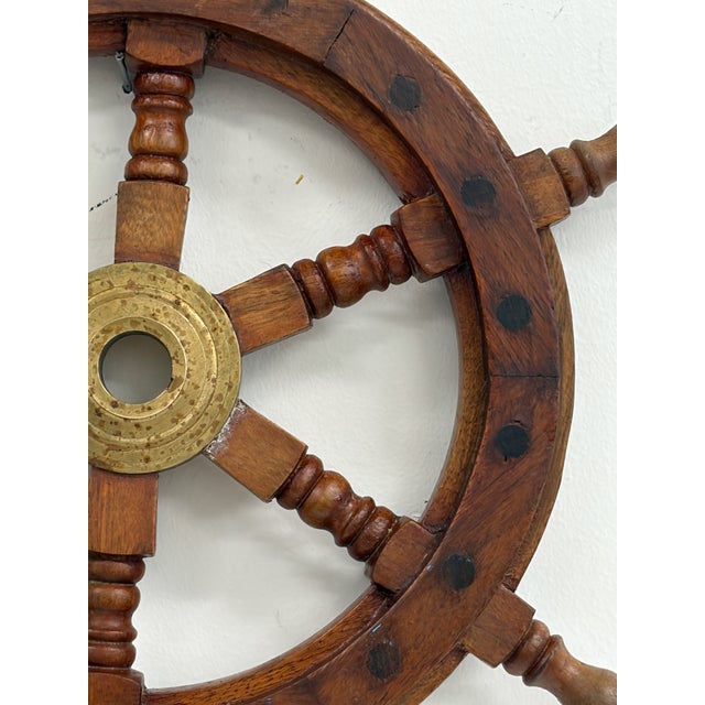 Small decorative ship’s wheel made of stained wood with turned handles and brass-tone center hub. Classic nautical design...