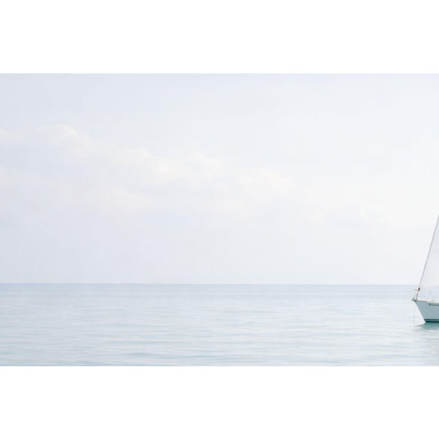 Minimalist coastal artwork featuring a single sailboat on calm water beneath a soft, muted sky. The composition emphasizes...