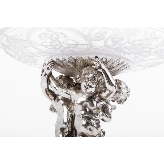Chiselled silver centerpiece topped with an engraved crystal cup carried by cherubs standing on a round tripod base...