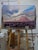 The painting depicts a vast desert landscape, featuring dramatic rock formations that rise majestically against a sky...