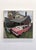 This oil on wood panel artwork captures a vibrant scene of a pink limousine resting in tall grass, juxtaposed against a...