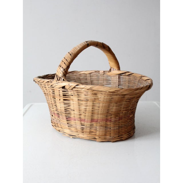 This is a vintage woven harvest basket with a wide, short handle and a soft pink stripe woven through the center. The...