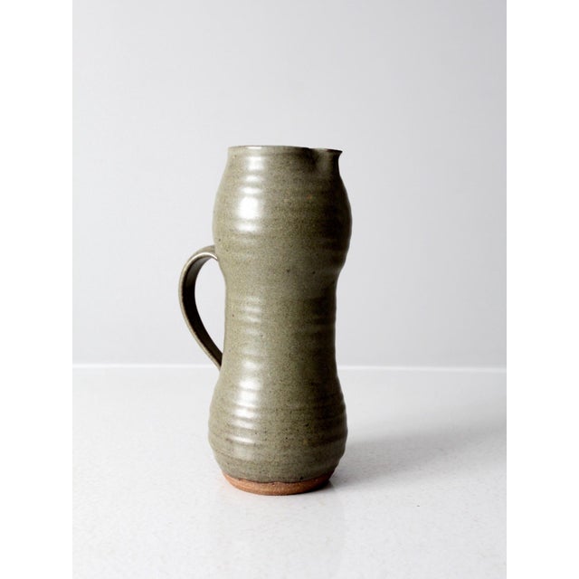 This vintage handmade studio pottery pitcher features a soft moss green glaze layered over a warm clay base, creating...