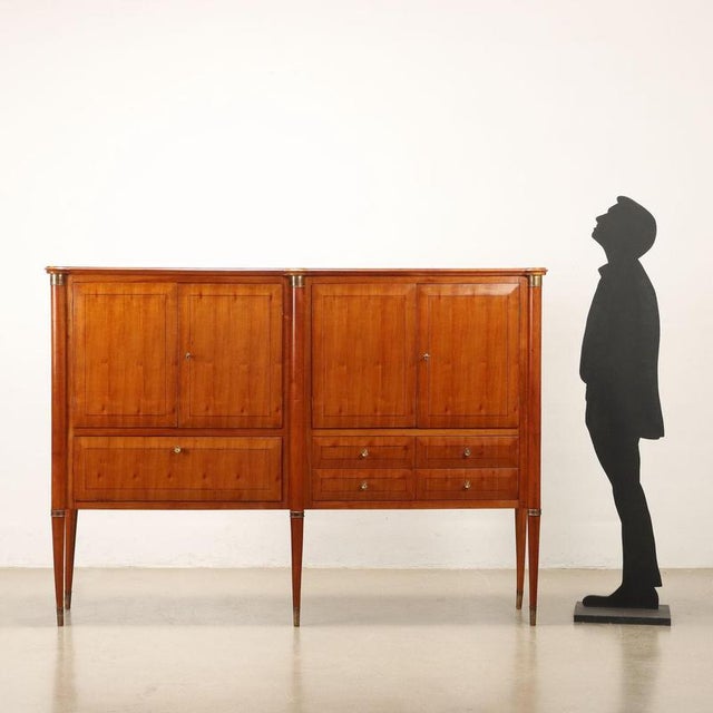 Elegant sideboard with hinged doors, flap and visible drawers. In cherry wood veneer with inlaid thread, brass details and...