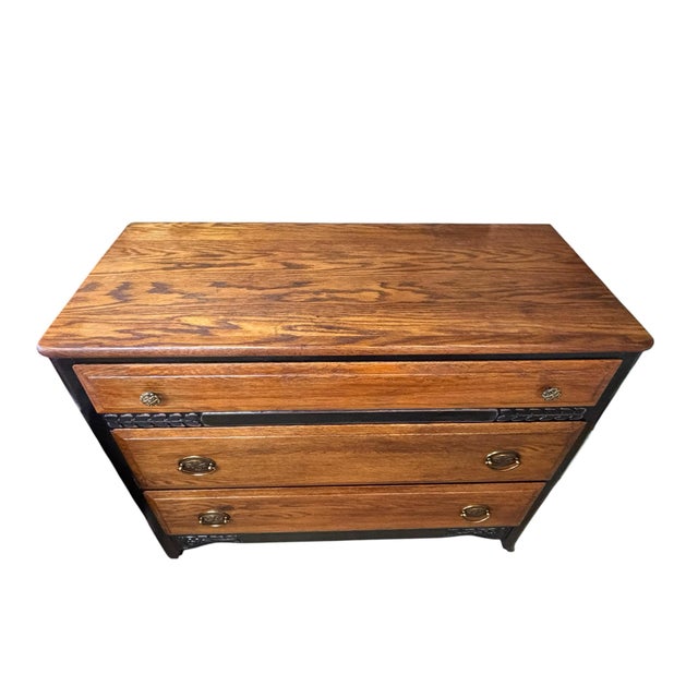 A distinctive vintage chest with warm oak drawer fronts set against a black-painted case. Carved foliate details between...