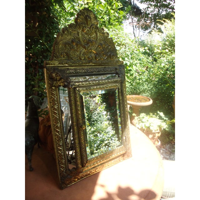 A small brush cabinet, richly decorated with embossed brass sheeting, the central mirror with a beveled edge, is...