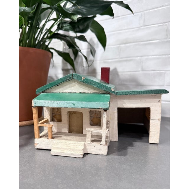 A handmade folk art model of a small house with porch, chimney, and attached garage, painted in white with a green roof...