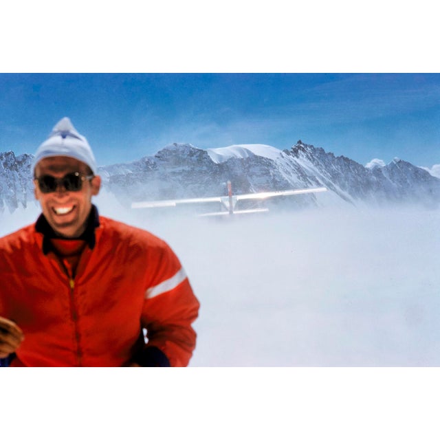 Haute Route Take Off by Toni Frissell A skier smiles from the top of a mountain in the clouds as a plane flies over head,...