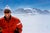 Haute Route Take Off by Toni Frissell A skier smiles from the top of a mountain in the clouds as a plane flies over head,...