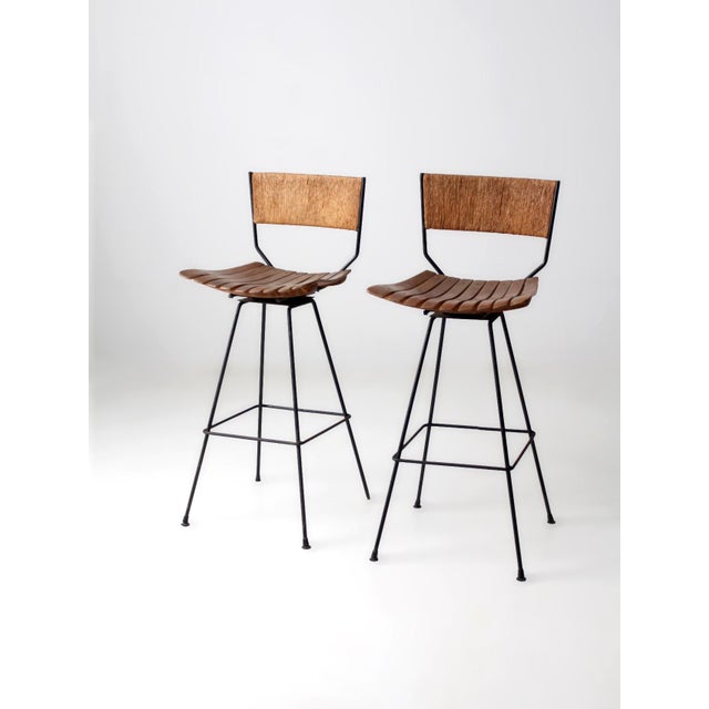 A pair of mid-century Arthur Umanoff style stools featuring black metal frames with woven rush backs and slatted wood...