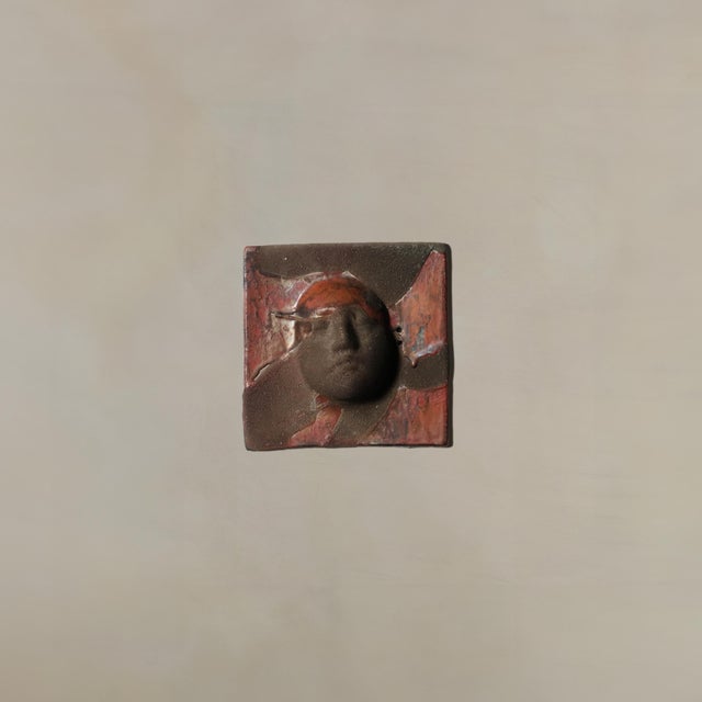 This handmade ceramic wall sculpture fuses abstract form and textured detail, centering on a sculpted visage that emerges...