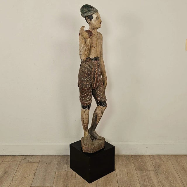 A 19th-century Southeast Asian tall painted and carved wood figure of a man, with wear and losses, on later stand. The man...