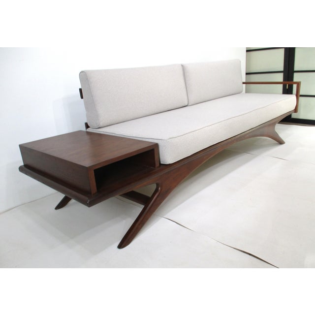 A unique sculptural walnut framed sofa daybed with splayed legs, built in end table with open storage to both sides and a...
