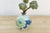 A small vintage vase with a pair of layered blossoms, one in soft green, one in deeper blue, share the body in a single...