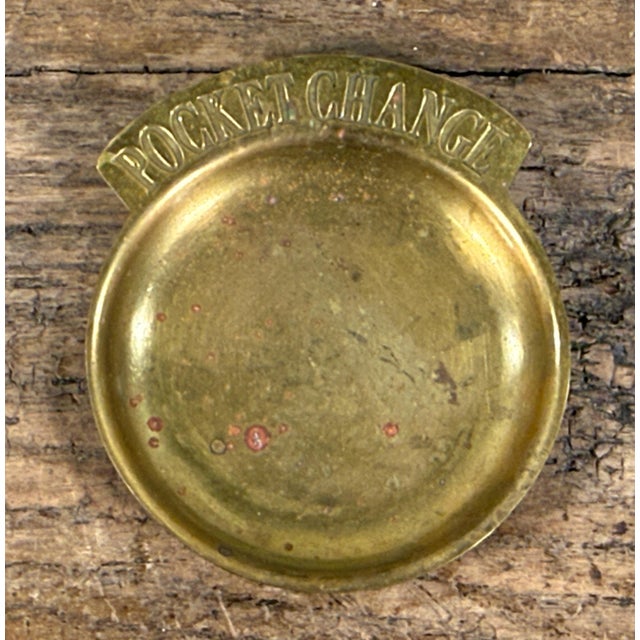 This brass catchall dish features a shallow oval form with a protruding tab stamped with the words "POCKET CHANGE" in...